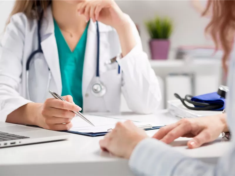Ein Arzt und eine Patientin sitzen an einem Tisch. Der Arzt macht Notizen und die Patientin hört aufmerksam zu.