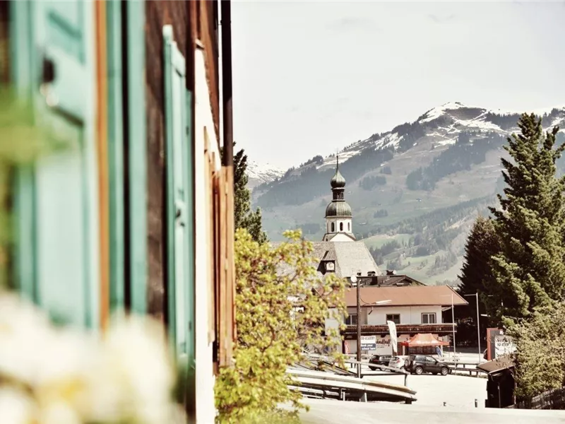 Blick auf eine Dorfkirche mit Zwiebelturm vor schneebedeckten Bergen, eingerahmt von einer Hausfassade.