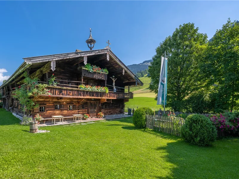 Ein traditionelles Holzhaus umgeben von saftigem Grün und Bäumen. Der Himmel ist klar und sonnig, was eine idyllische Berglandschaft schafft.