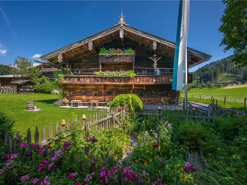 Ein traditionelles Alpenhaus umgeben von einem schönen Garten. Die Fassade ist mit Blumen geschmückt und der Himmel ist klar und blau.