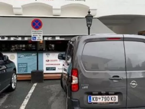 Ein graues Fahrzeug steht in einer Parklücke vor einem Gebäude mit dem Schriftzug "Schelhammers Capital 1832". Die Umgebung ist urban und aufgeräumt, mit weiteren Fahrzeugen und Straßenschildern in der Nähe.