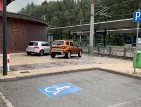 Ein Parkplatz mit zwei Autos, eines davon auf einem behindertengerechten Platz. Im Hintergrund ist ein Bahngleis und ein Gebäude sichtbar.