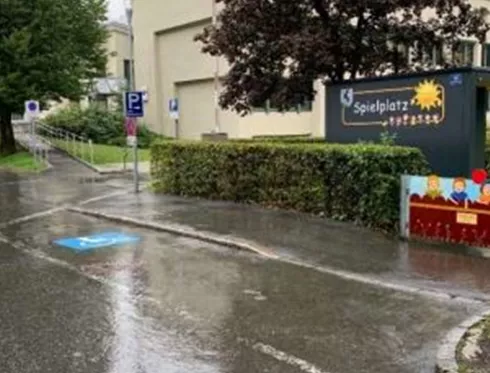Eine nasse Straße mit einem Spielplatzschild und Bäumen im Hintergrund. Es regnet leicht, und es sind keine Menschen zu sehen.