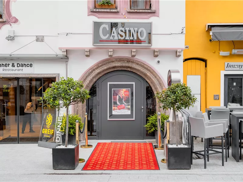 Ein Casino-Eingang mit einem roten Teppich und grünen Pflanzen. Daneben befindet sich ein Restaurant und weitere Sitzgelegenheiten.
