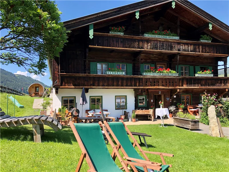 Ein traditionelles Holzhaus umgeben von einer grünen Wiese. Liegestühle laden zum Entspannen ein, und die Berglandschaft ist im Hintergrund sichtbar.