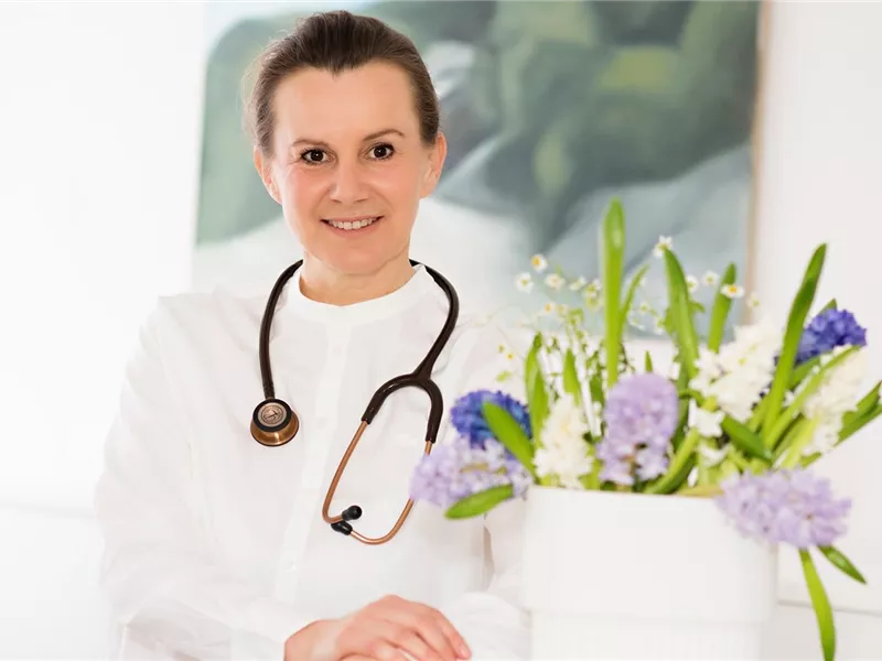 Eine lächelnde Ärztin in weißem Kittel steht vor einem Blumenarrangement. Sie trägt ein Stethoskop um den Hals und strahlt Professionalität aus.