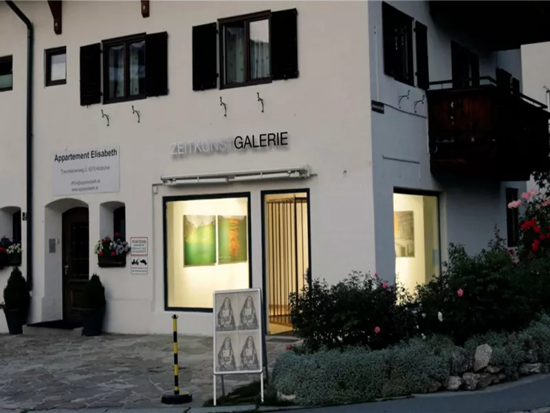 Ein modernes Galeriegebäude mit beleuchteten Fenstern. Vor dem Eingang sind Blumen und ein paar Personen zu sehen.