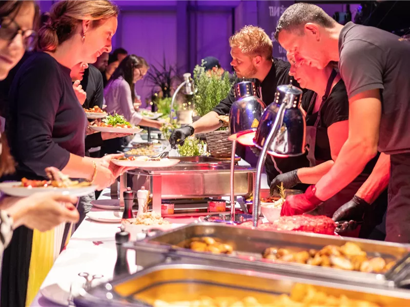 Eine lebhafte Buffet-Szene mit mehreren Personen, die Essen servieren und auswählen. Der Tisch ist mit verschiedenen Gerichten und frischen Zutaten dekoriert.