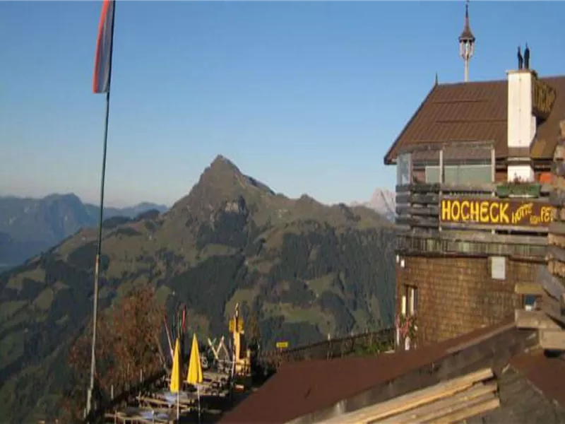 Ein Bergpanorama mit einer Berghütte und einer Fahne im Vordergrund. Im Hintergrund sind sanfte Hügel und ein klarer Himmel zu sehen.