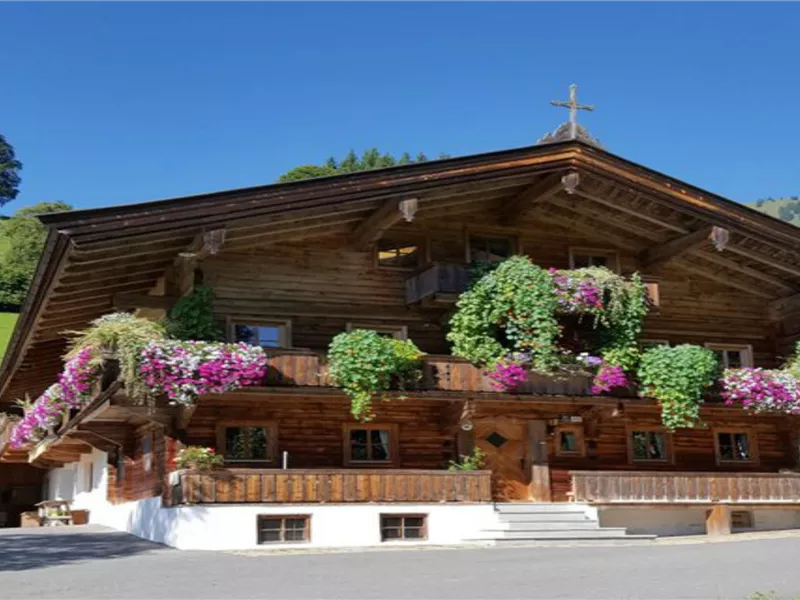 Ein traditionelles Holzhaus mit bunten Blumen auf dem Balkon. Im Hintergrund sind grüne Hügel und ein klarer blauer Himmel zu sehen.