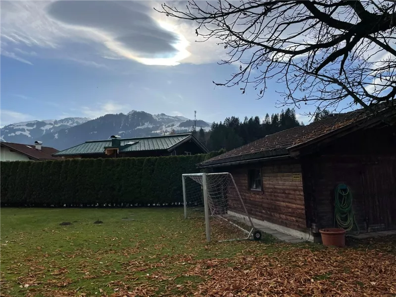 Ein schöner Garten mit einem Holzhaus und einem Fußballtor. Im Hintergrund sind Berge und ein bewölkter Himmel zu sehen.