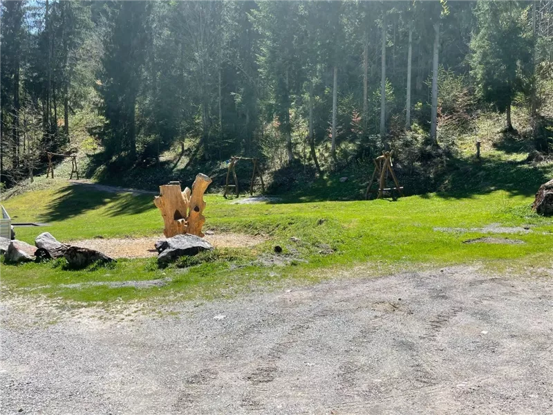 Ein schöner grüner Bereich mit Holzskulpturen und Bäumen im Hintergrund. Der Boden ist teilweise mit Kies bedeckt und es gibt viel Sonnenlicht.