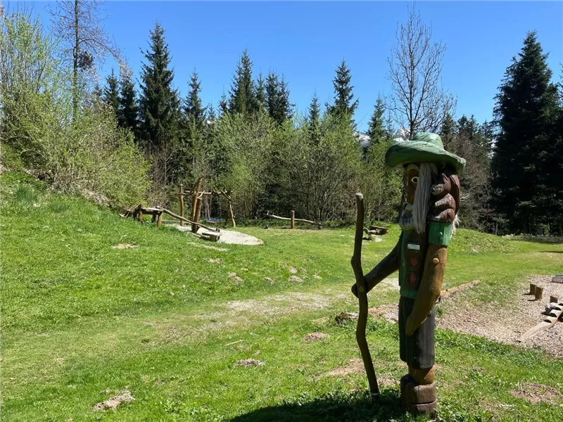 Eine hölzerne Skulptur eines Wanderers mit einem Stock steht auf einer grünen Wiese. Im Hintergrund sind Bäume und Spielgeräte sichtbar.