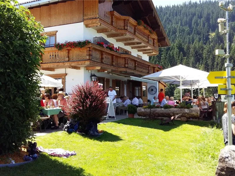 Ein charmantes Alpenhaus mit einem schönen Außenbereich. Menschen genießen die Sonne und einen Blick auf die umliegenden Berge.