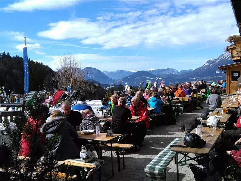 Eine lebhafte Bergterrasse mit vielen Gästen, die bei schönem Wetter entspannen. Im Hintergrund sind schneebedeckte Berge und ein blauer Himmel zu sehen.