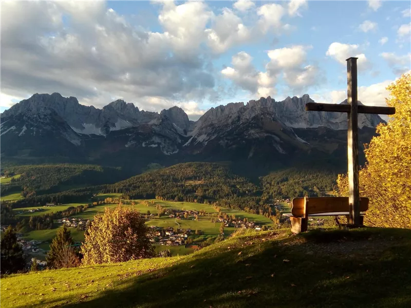 Eine malerische Berglandschaft mit majestätischen Gipfeln und einem bordeauxfarbenen Kreuz im Vordergrund. Die Szenerie zeigt eine ruhige und friedliche Natur im goldenen Licht des Sonnenuntergangs.