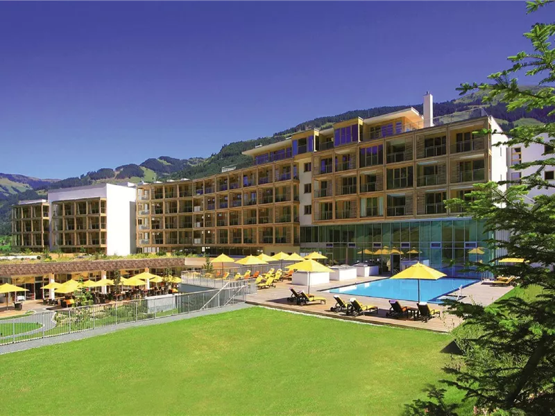 Ein modernes Hotel mit einem schönen Poolbereich und Liegestühlen. Im Hintergrund sind Berge und ein klarer Himmel zu sehen.