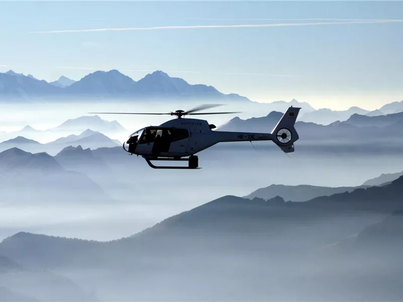 Ein Hubschrauber schwebt über einem nebligen Bergpanorama. Die umliegenden Gipfel sind in sanften Blautönen getaucht.