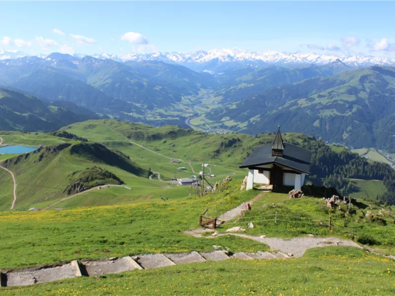 Eine malerische Berglandschaft mit saftigem Grün und schneebedeckten Gipfeln im Hintergrund. Im Vordergrund steht eine kleine Kapelle und ein Steinweg führt hinunter.