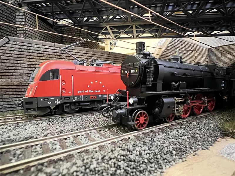 Zwei Züge stehen auf einem Gleis in einer Modellanlage. Der rote Zug ist modern, während die schwarze Lokomotive klassisch aussieht.