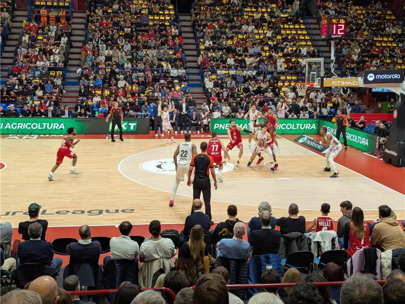 Ein Basketballspiel in einem vollen Stadion mit begeisterten Zuschauern. Zwei Mannschaften kämpfen um den Ball auf dem Spielfeld.