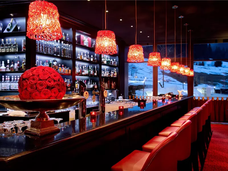 Eine stilvolle Bar mit roten Lampenschirmen und einer eleganten Einrichtung. Im Hintergrund sind schneebedeckte Landschaften zu sehen.