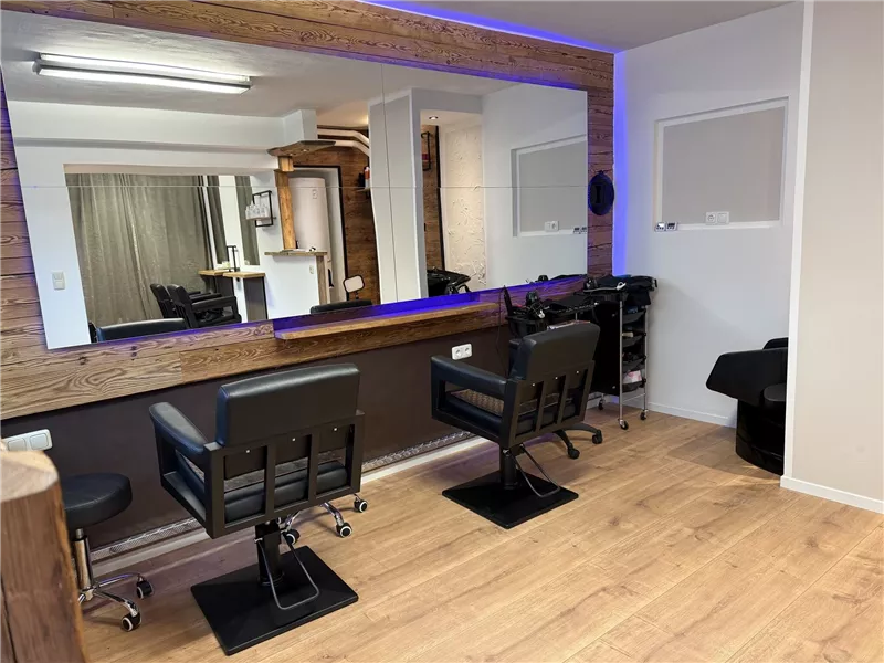 Ein modernes Friseurstudio mit zwei schwarzen Stühlen und einem großen Spiegel. Der Raum hat einen warmen Holzboden und eine gemütliche Atmosphäre.