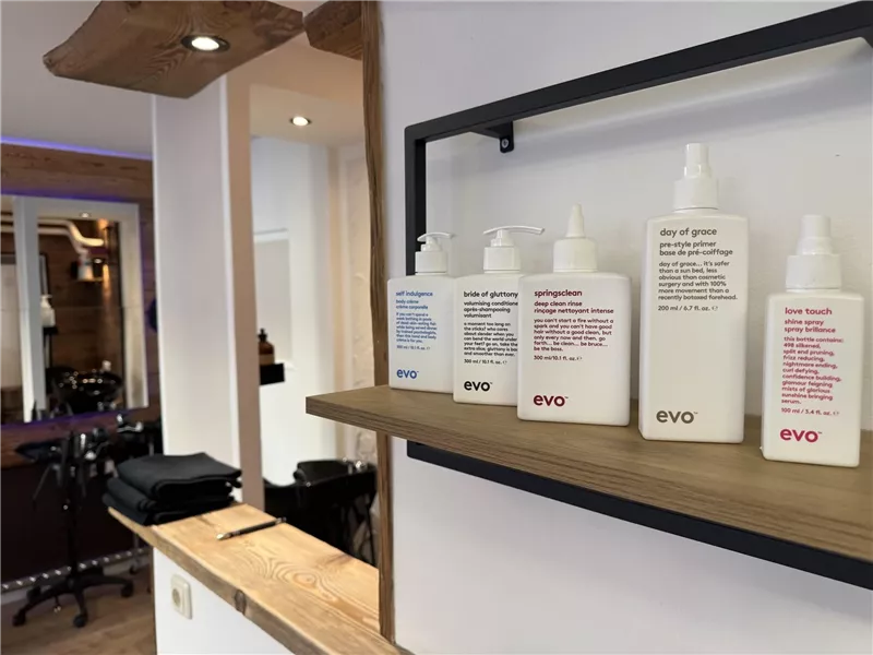Auf einem Holzregal stehen verschiedene Flaschen mit Haarpflegeprodukten der Marke Evo. Der Hintergrund zeigt einen Friseursalon mit dunklem Mobiliar.