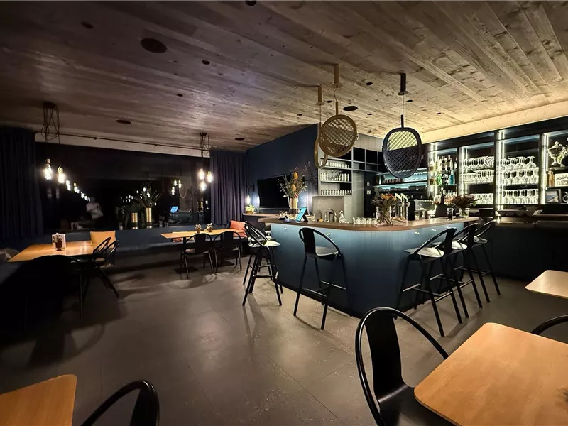 Ein modernes Restaurant mit einer stilvollen Bar und Holzelementen. Die gemütliche Atmosphäre wird durch sanfte Beleuchtung und dunkle Farben geprägt.