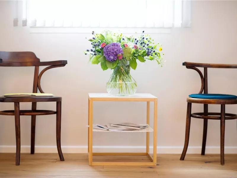 Zwei Holzstühle stehen neben einem kleinen Tisch. Auf dem Tisch befindet sich eine Vase mit bunten Blumen.