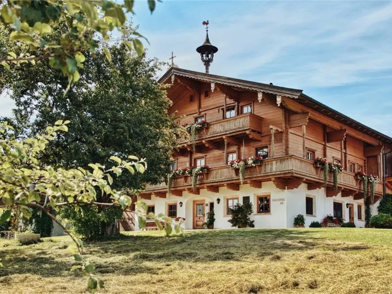 Ein traditionelles Holzhaus mit Balkonen und bunten Blumen. Im Vordergrund steht ein Baum und der Himmel ist klar.