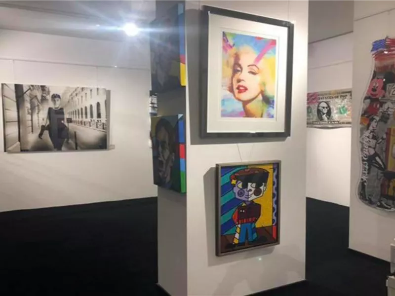 Eine Kunstgalerie mit mehreren Rahmen und bunten Gemälden. Die Wände sind mit verschiedenen Kunstwerken dekoriert, darunter ein Porträt und eine Fotografie.