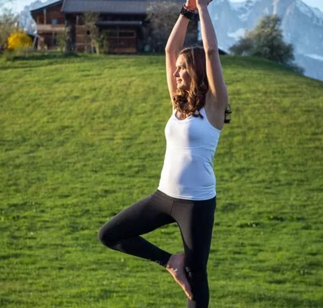 Eine Frau praktiziert Yoga in der Baum-Position auf einem Felsen vor einer Bergkulisse.