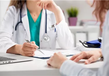 Eine Ärztin spricht mit einer Patientin in einem medizinischen Raum. Auf dem Tisch liegen Dokumente und ein Laptop.