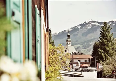 Blick auf eine Dorfkirche mit Zwiebelturm vor schneebedeckten Bergen, eingerahmt von einer Hausfassade.