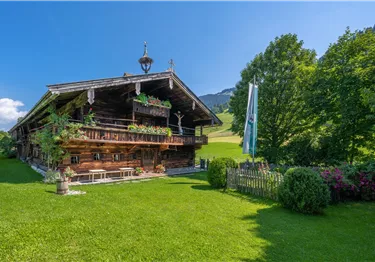 Ein traditionelles Holzhaus umgeben von saftigem Grün und Bäumen. Der Himmel ist klar und sonnig, was eine idyllische Berglandschaft schafft.