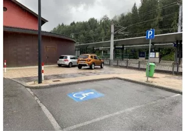 Ein Parkplatz mit zwei Autos, eines davon auf einem behindertengerechten Platz. Im Hintergrund ist ein Bahngleis und ein Gebäude sichtbar.