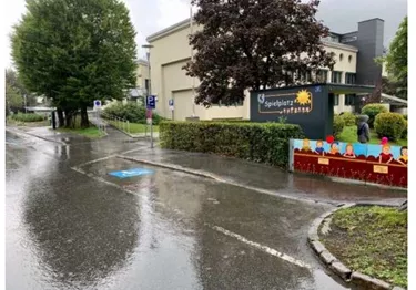Eine nasse Straße mit einem Spielplatzschild und Bäumen im Hintergrund. Es regnet leicht, und es sind keine Menschen zu sehen.