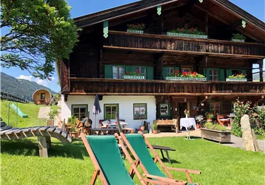 Ein traditionelles Holzhaus umgeben von einer grünen Wiese. Liegestühle laden zum Entspannen ein, und die Berglandschaft ist im Hintergrund sichtbar.