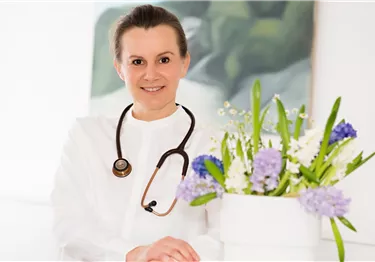 Eine lächelnde Ärztin in weißem Kittel steht vor einem Blumenarrangement. Sie trägt ein Stethoskop um den Hals und strahlt Professionalität aus.