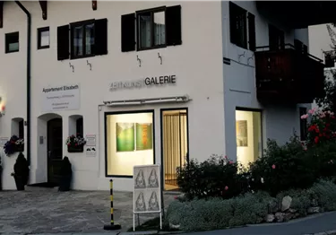 Ein modernes Galeriegebäude mit beleuchteten Fenstern. Vor dem Eingang sind Blumen und ein paar Personen zu sehen.