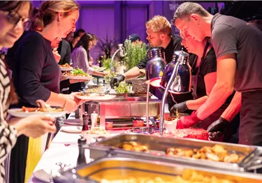 Eine lebhafte Buffet-Szene mit mehreren Personen, die Essen servieren und auswählen. Der Tisch ist mit verschiedenen Gerichten und frischen Zutaten dekoriert.