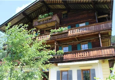 Ein traditionelles Holzhaus mit mehreren Balkonen und grünen Pflanzen. Im Hintergrund ist eine schöne, natürliche Umgebung zu sehen.