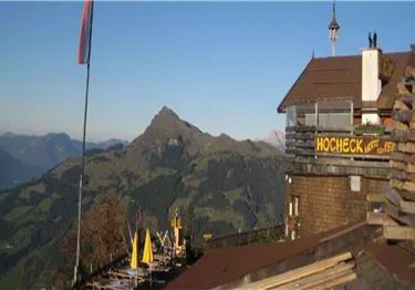 Ein Bergpanorama mit einer Berghütte und einer Fahne im Vordergrund. Im Hintergrund sind sanfte Hügel und ein klarer Himmel zu sehen.