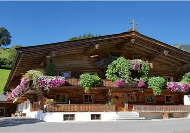 Ein traditionelles Holzhaus mit bunten Blumen auf dem Balkon. Im Hintergrund sind grüne Hügel und ein klarer blauer Himmel zu sehen.