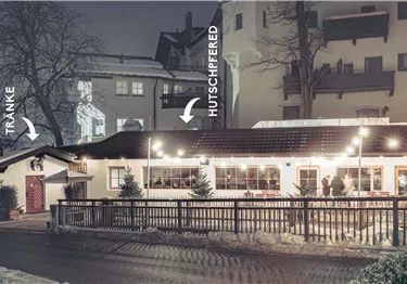 Ein gemütliches Restaurant im Winter, beleuchtet mit warmen Lichtern. Schnee liegt auf dem Boden und die Umgebung wirkt ruhig und einladend.