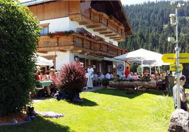 Ein charmantes Alpenhaus mit einem schönen Außenbereich. Menschen genießen die Sonne und einen Blick auf die umliegenden Berge.
