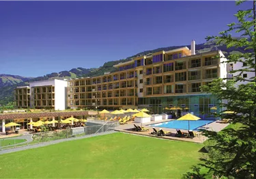 Ein modernes Hotel mit einem schönen Poolbereich und Liegestühlen. Im Hintergrund sind Berge und ein klarer Himmel zu sehen.