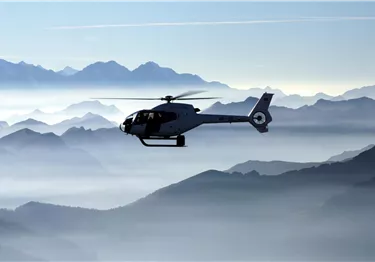 Ein Hubschrauber schwebt über einem nebligen Bergpanorama. Die umliegenden Gipfel sind in sanften Blautönen getaucht.