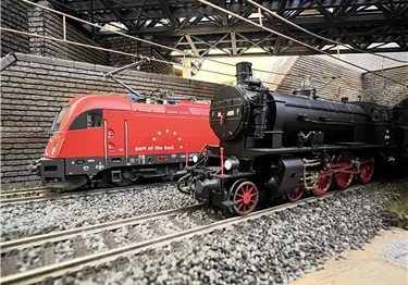 Zwei Züge stehen auf einem Gleis in einer Modellanlage. Der rote Zug ist modern, während die schwarze Lokomotive klassisch aussieht.